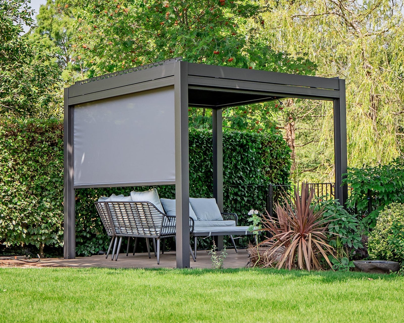 Anthracite grey louvered pergola with a lowered grey side blind and rattan lounge seating on a wood deck in a green residential garden.