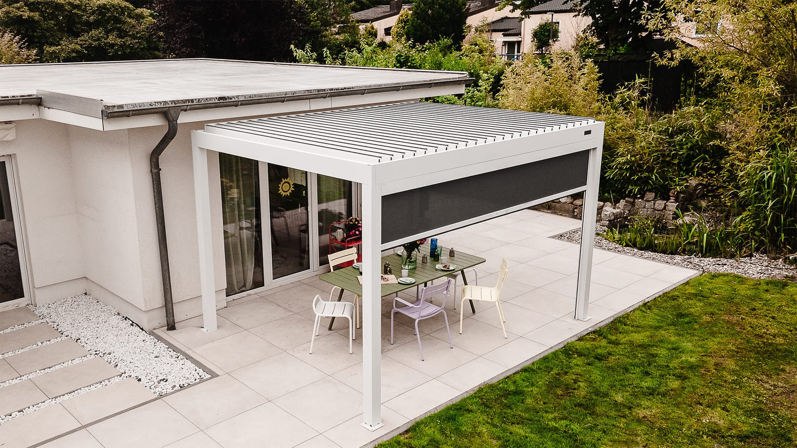 White louvered-roof pergola with a side blind screen attached to a modern house over a tiled patio with a green dining table and chairs.