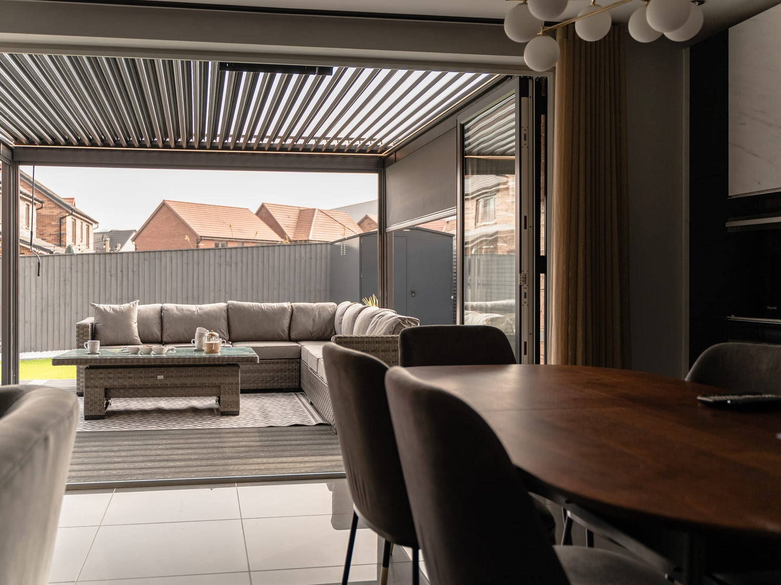 Anthracite grey louvered pergola viewed from interior dining room through open bifold doors, with rattan corner sofa and coffee table on patio, daytime.