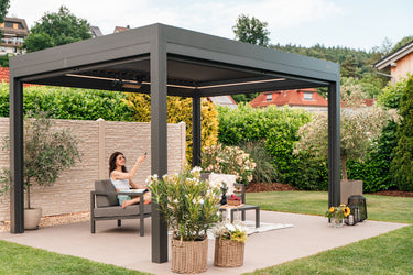 Woman sitting under a anthracite pergola in a garden setting with plants and furniture.