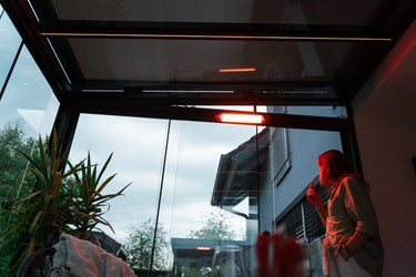 Person standing by a glass door inside a conservatory with a heater inside, reflecting on a cloudy day.