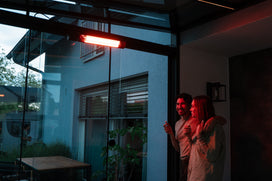 Two people standing by a glass door with a heater inside a conservatory, looking outside.