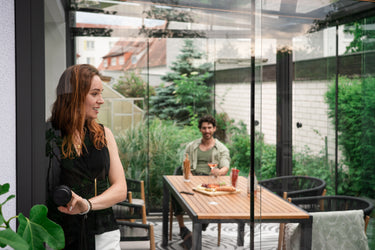 Woman standing in a modern conservatory with a man sitting at a table in the background.