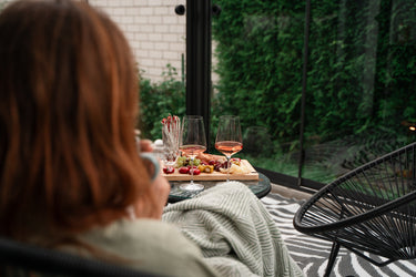 Person sitting in a conservatory with a glass of wine and a platter of food, looking out at greenery.